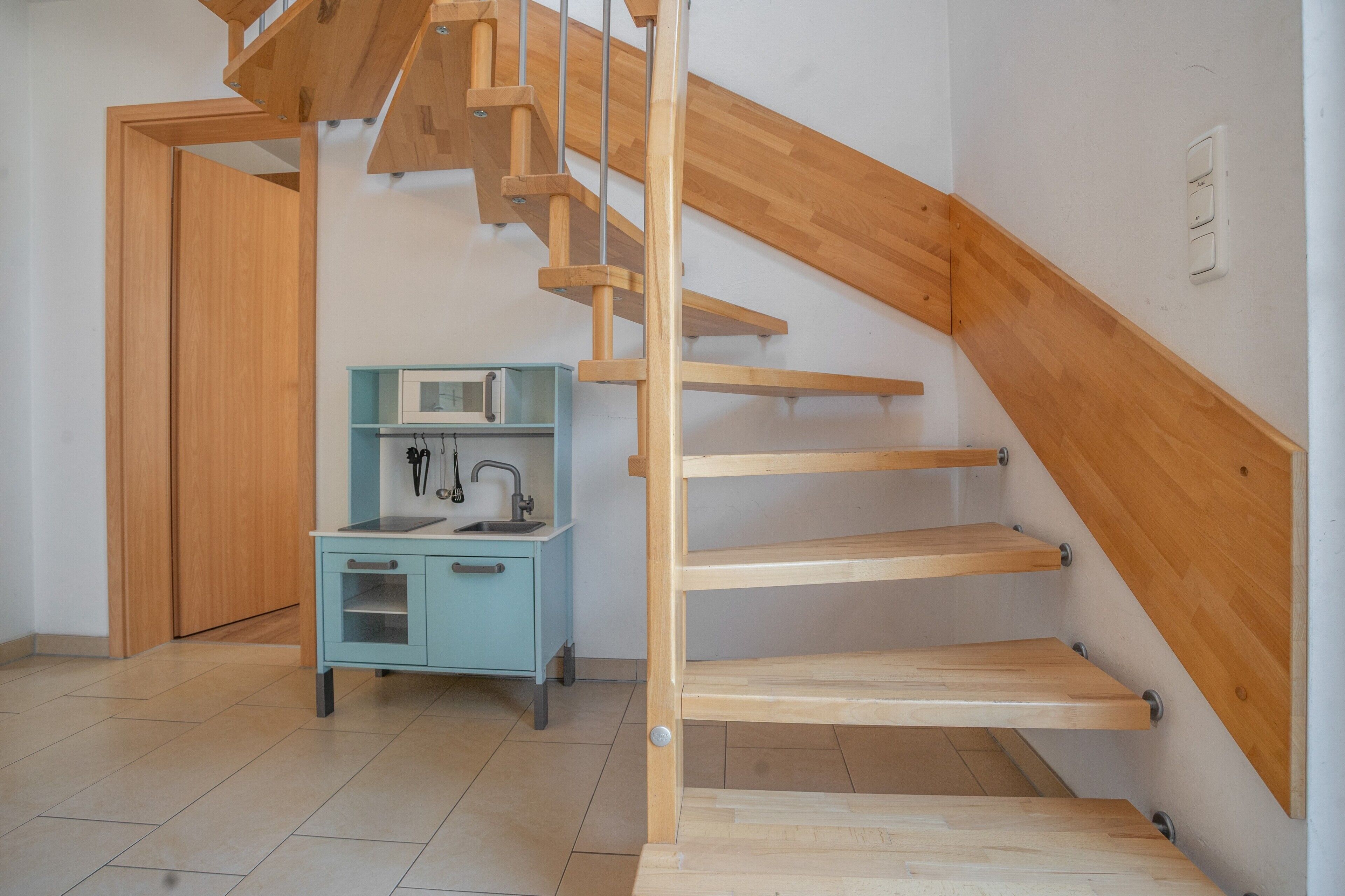 Een kamer met een houten trap, een kitchenette met een spoelbak en kastjes, en een deur naar een andere kamer.