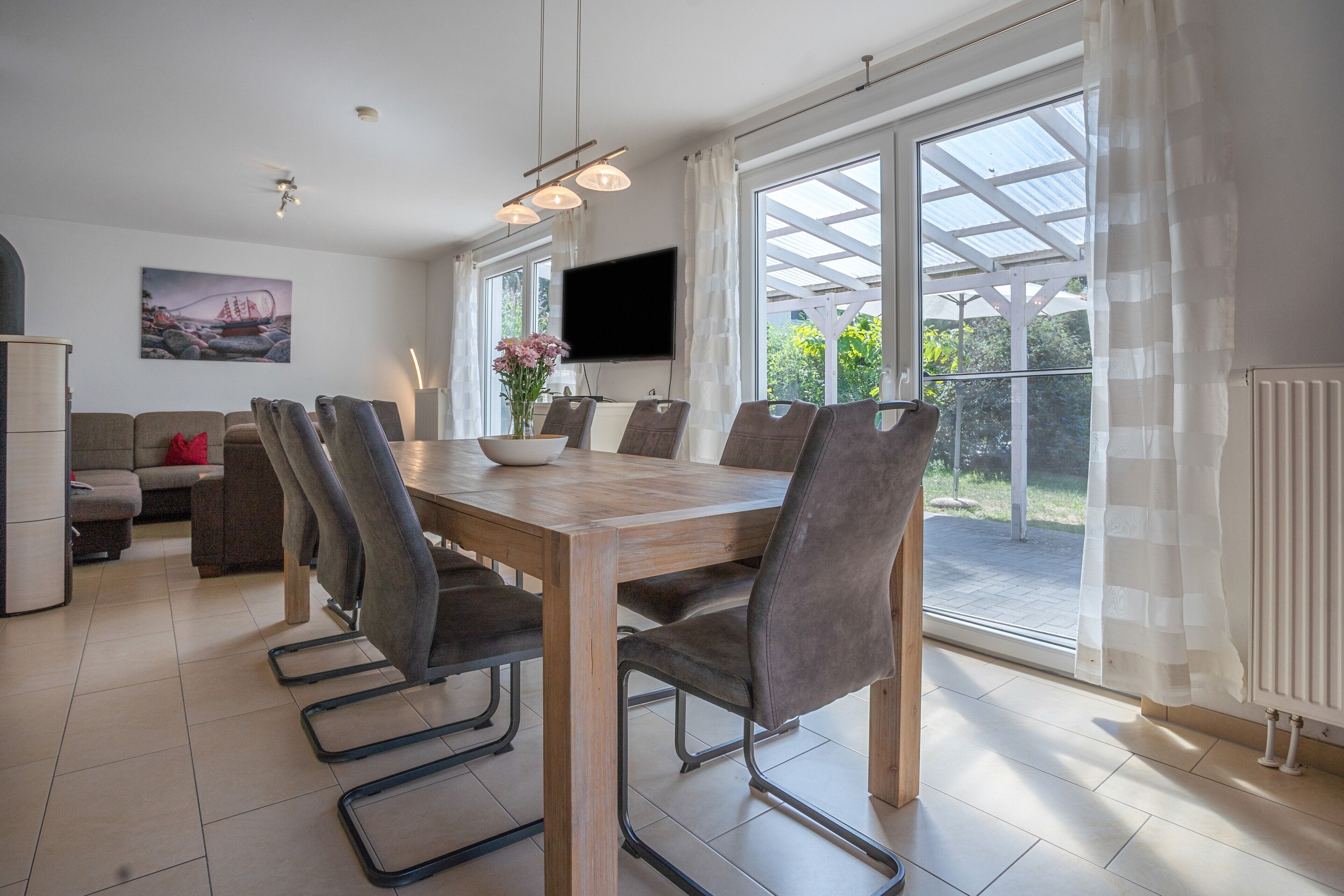 A dining area with a wooden table and chairs, a flat-screen TV mounted on the wall, and a view of the outdoors through large windows.