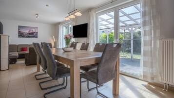 A dining area with a wooden table and chairs, a flat-screen TV mounted on the wall, and a view of the outdoors through large windows.