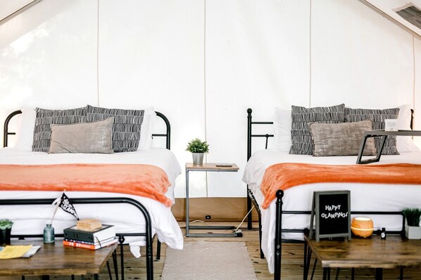 A room with two beds, each with a black metal frame, white bedding, and striped pillows. There is a small table with a plant, a sign that says 'HAPPY CAMPING,' and a wooden floor.