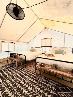 Ein Zeltraum mit einem Bett, einem kleinen Tisch, einer Bank und einem an der Decke befestigten Ventilator.
