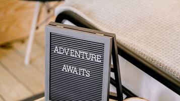 A bed with a headboard, a bedside table with a sign that reads 'ADVENTURE AWAITS,' a wooden candle holder, and a small card on the table.