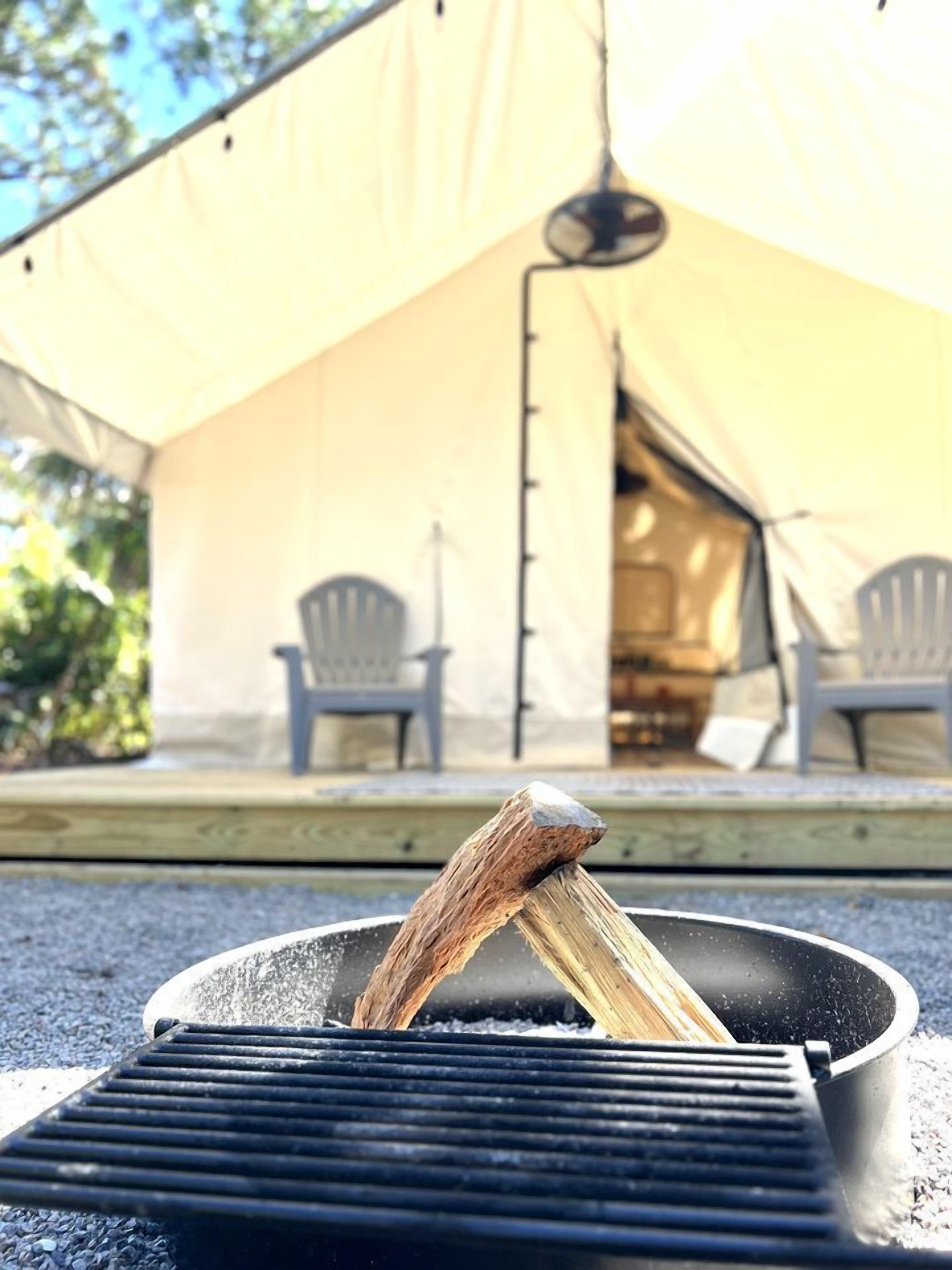 A grill with a stick over it, a tent, and two chairs in the background.