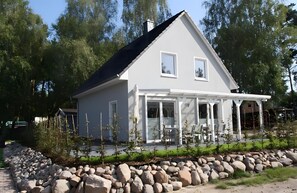 Ein modernes, zweistöckiges Haus mit überdachter Veranda, einer Steinmauer und einem Garten mit jungen Bäumen.