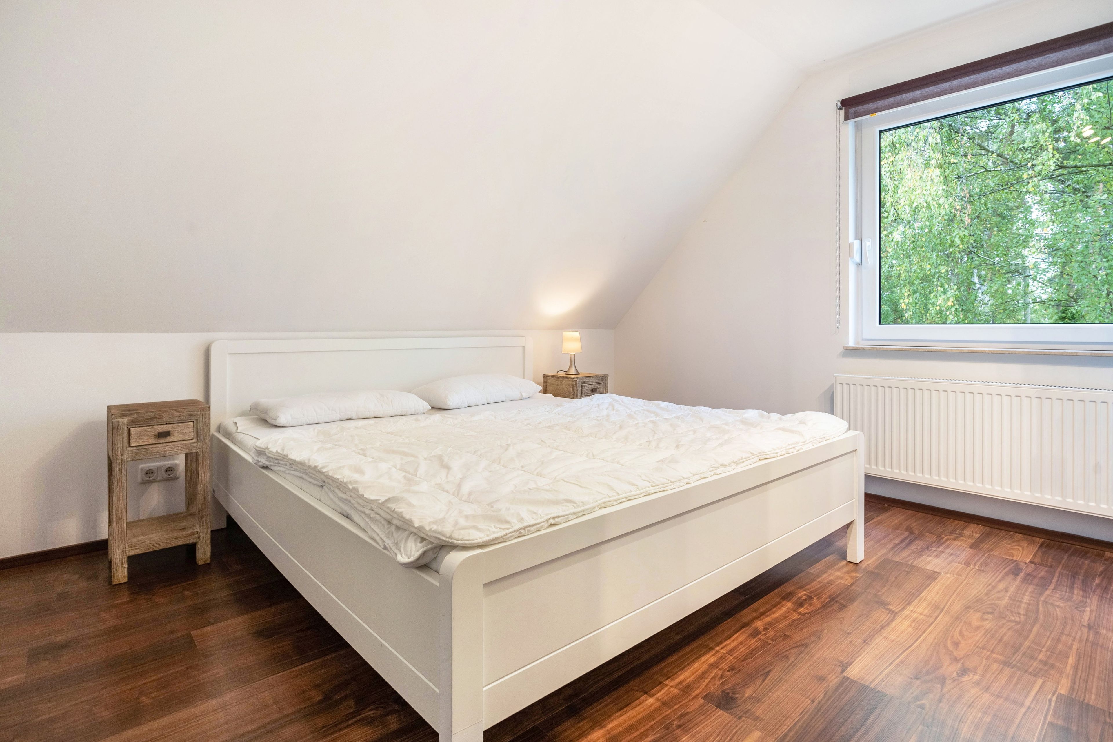A bedroom with a white bed, a nightstand, a lamp, and a window with a view of trees.