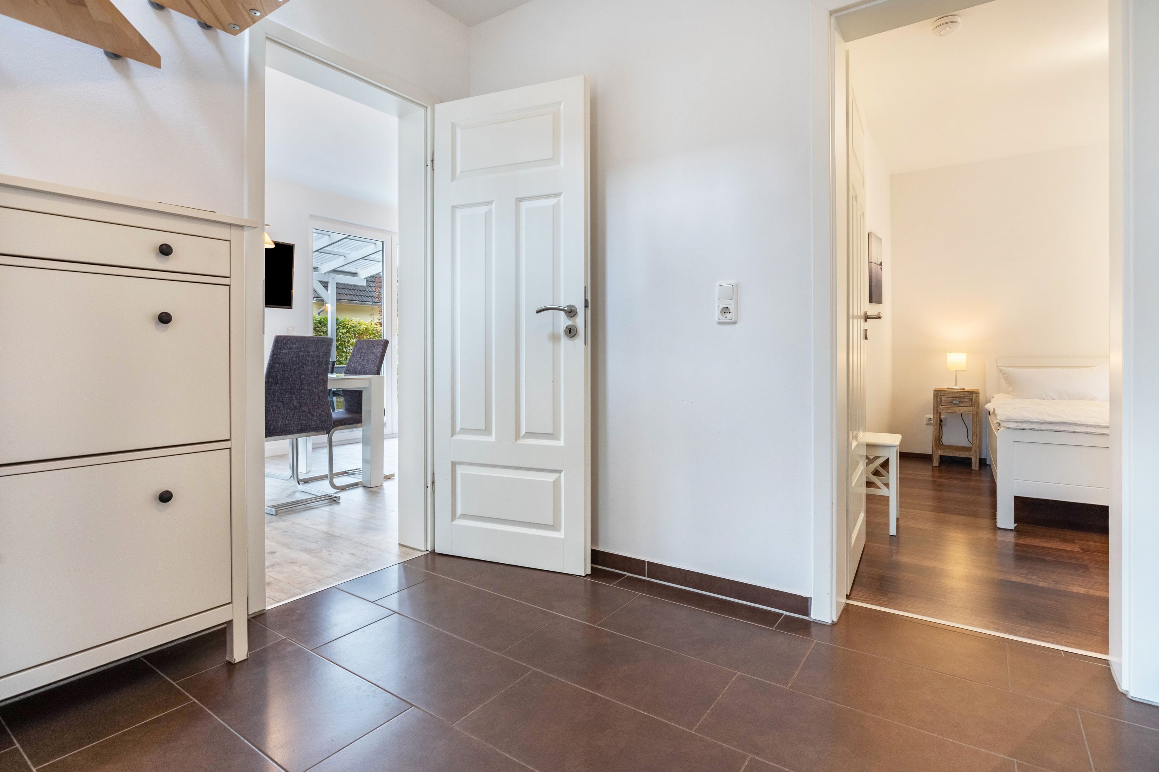 Habitación de hotel con cama, mesita de noche y un pequeño taburete. Se ve una puerta que da a un balcón con mobiliario exterior.