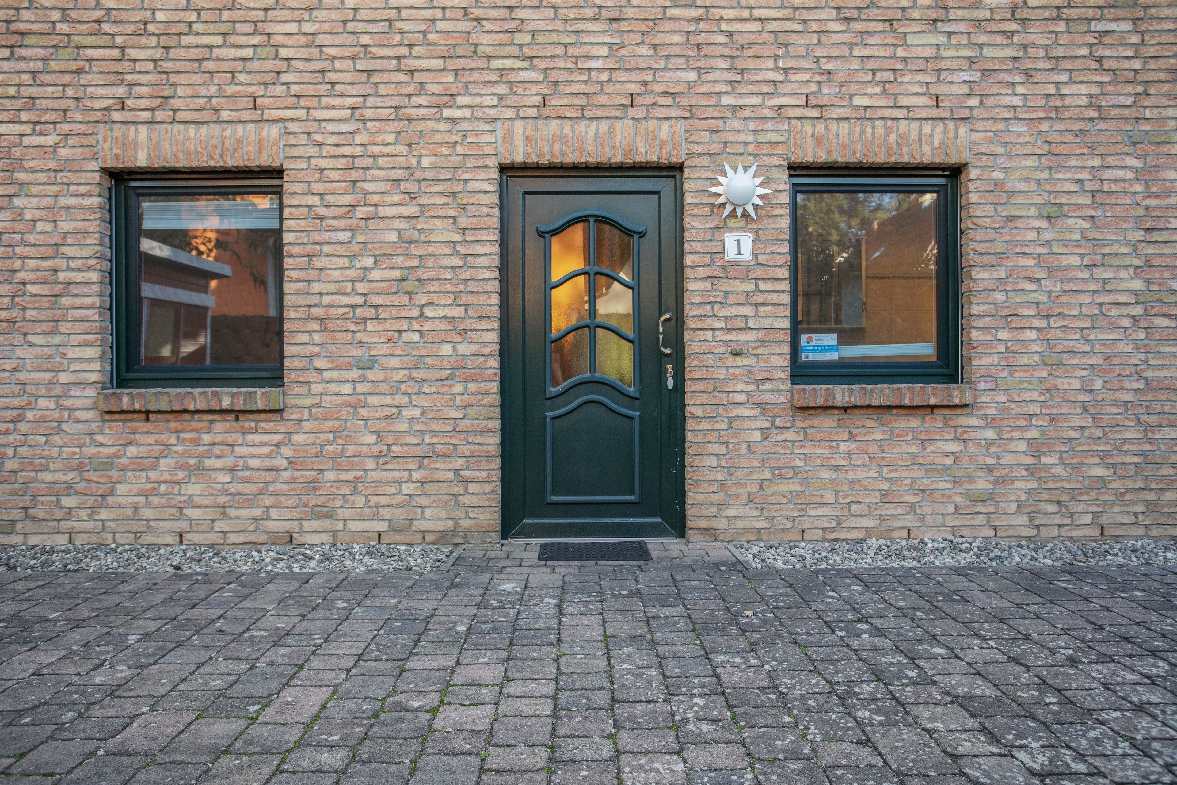 Een bakstenen gebouw met een donkergroene deur, twee ramen en een klein bord aan de muur.