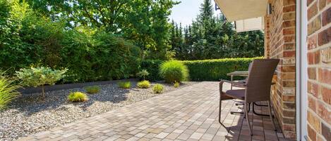 A patio with a brick floor, a table, and chairs, surrounded by greenery and trees.