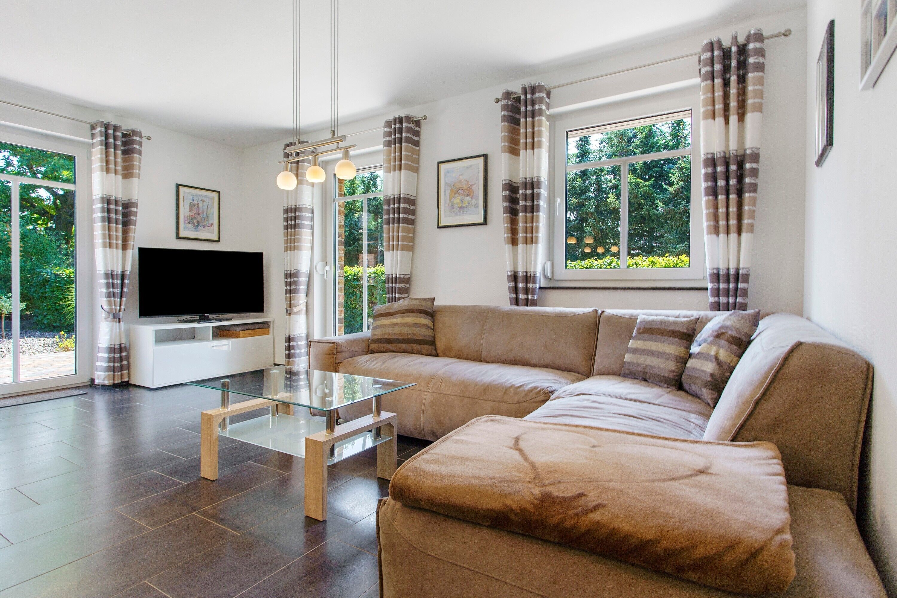 A modern living room with a sectional sofa, a glass coffee table, a flat-screen TV, and large windows with curtains.