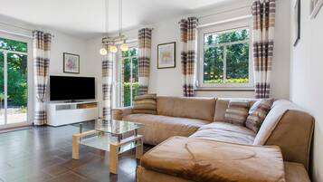 A modern living room with a sectional sofa, a glass coffee table, a flat-screen TV, and large windows with curtains.
