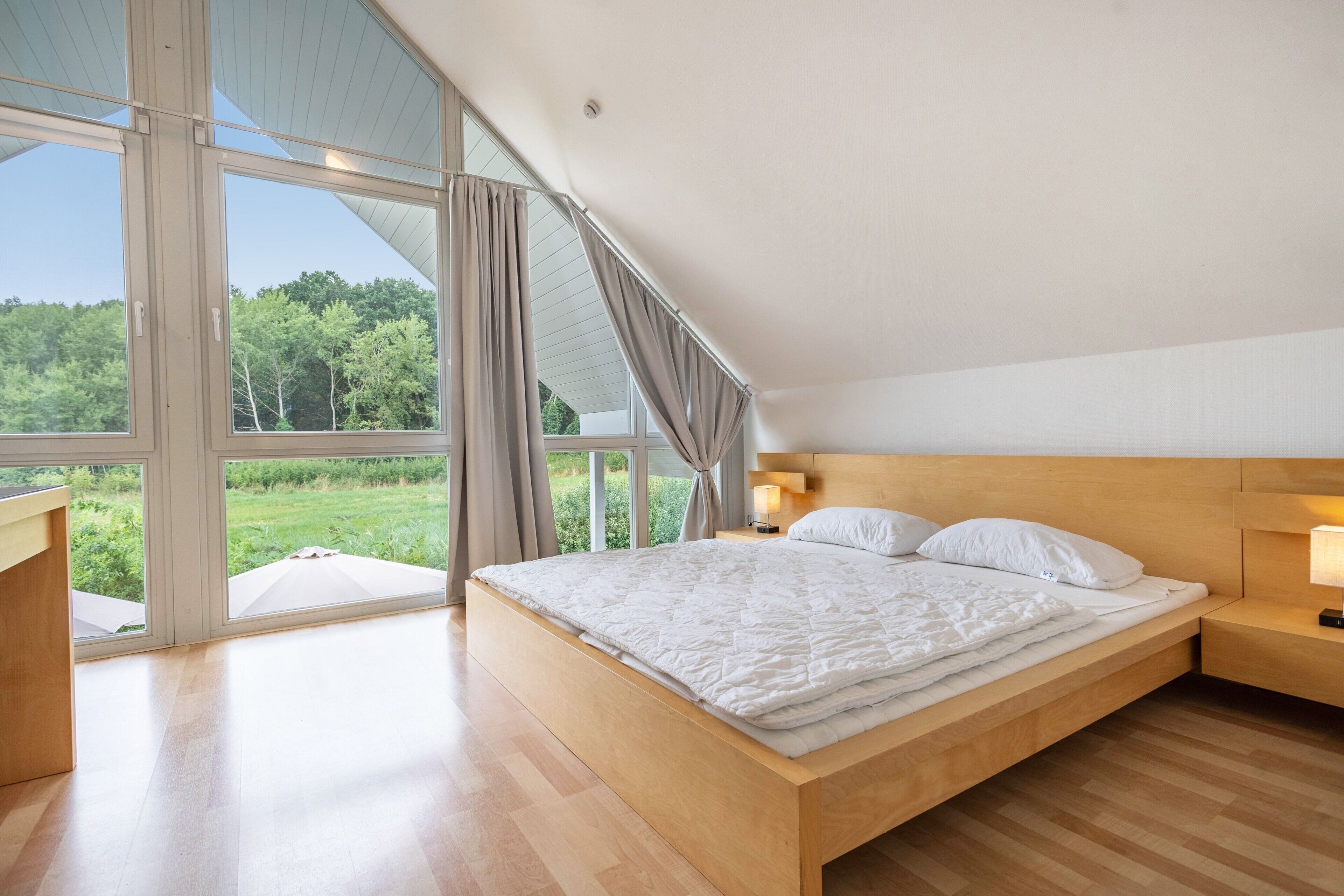 Une chambre à coucher avec un grand lit, une tête de lit en bois, des lampes de chevet et une vue sur la verdure grâce à de grandes fenêtres.