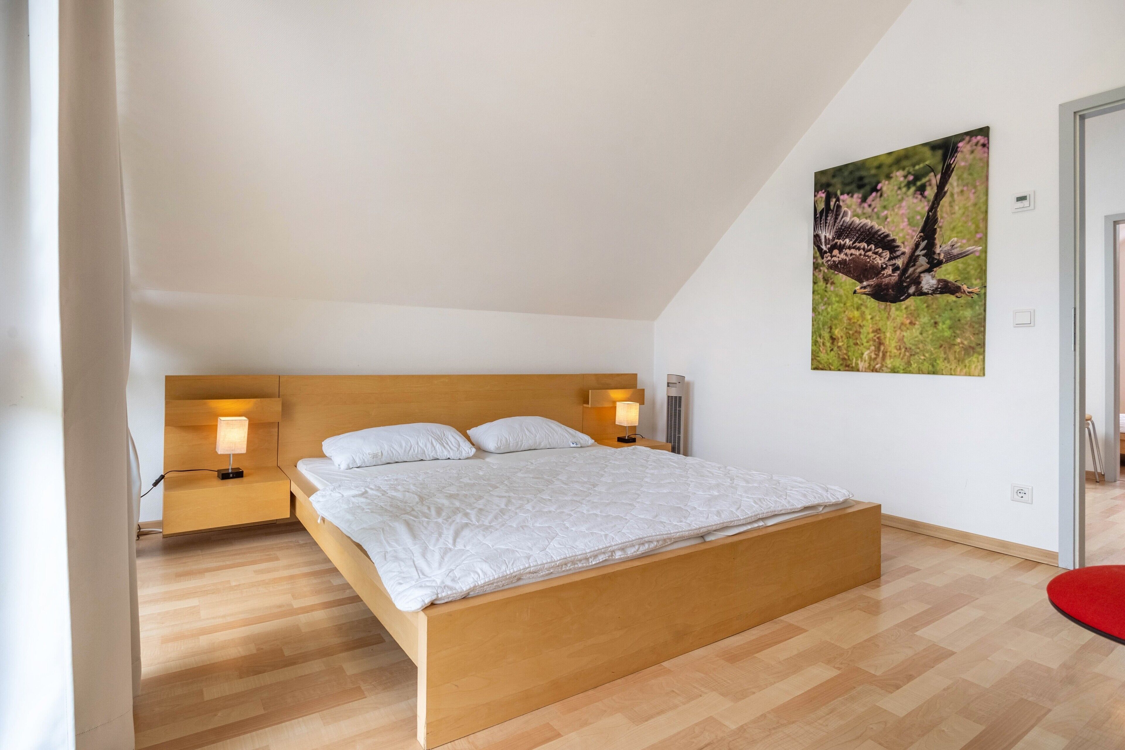 A bedroom with a wooden bed, two bedside tables with lamps, a wall-mounted air conditioner, and a framed picture of a bird.