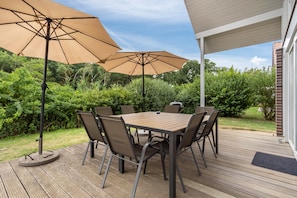 Een terras buiten met een houten dek, een ronde tafel, zes stoelen en twee grote parasols.