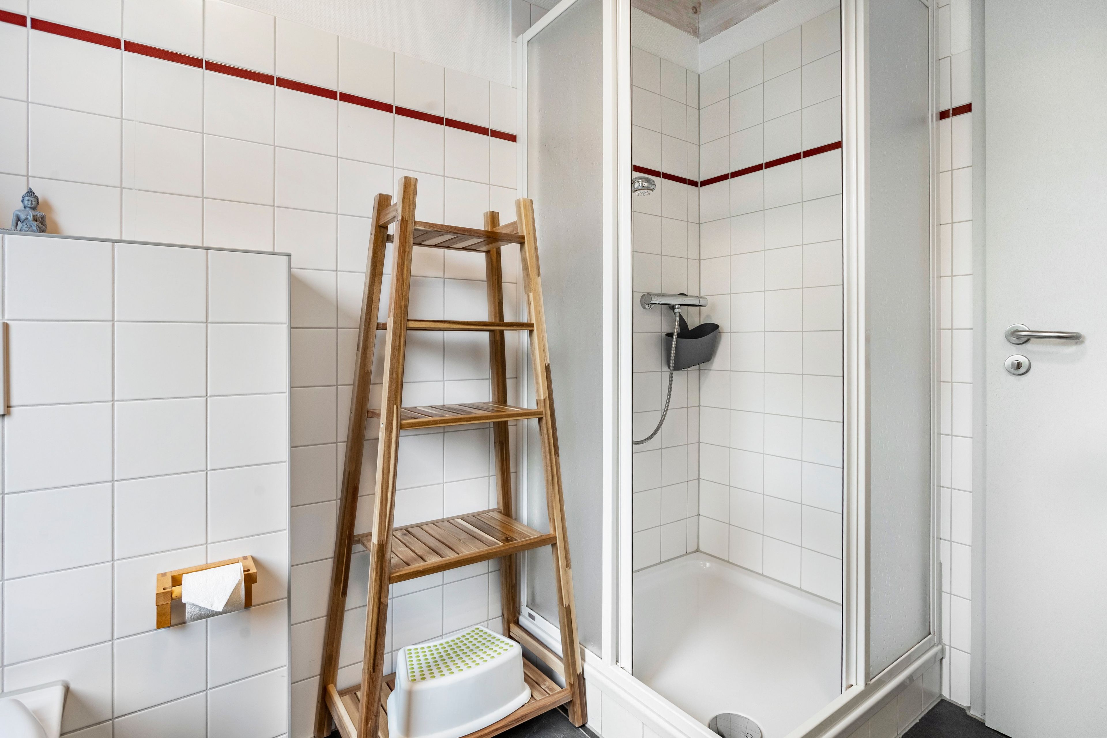 Une salle de bain avec des murs carrelés de carreaux blancs, un petit escabeau en bois, un espace douche et des toilettes.