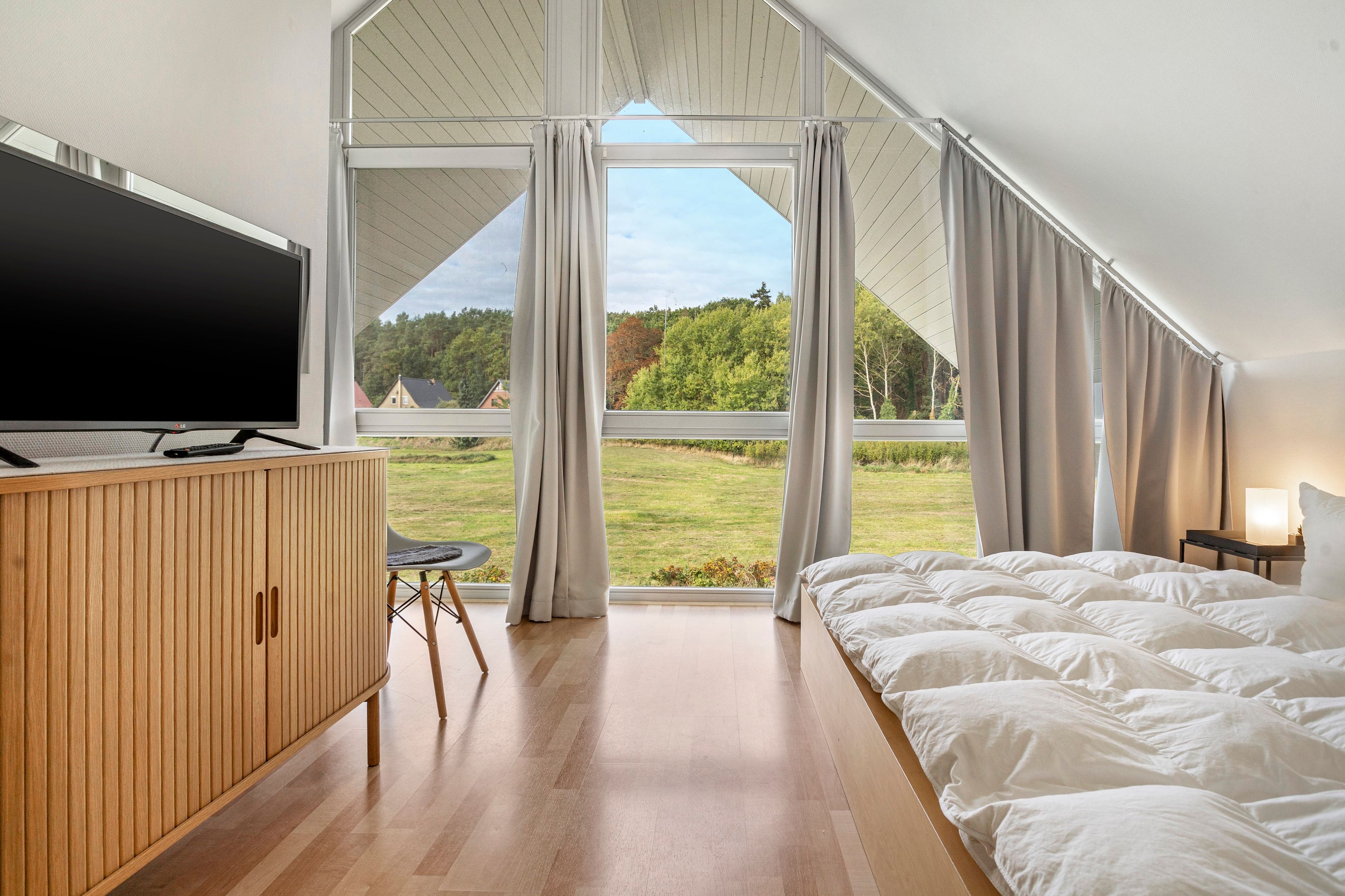 A bedroom with a bed, a TV, a desk, and a view of a green landscape.