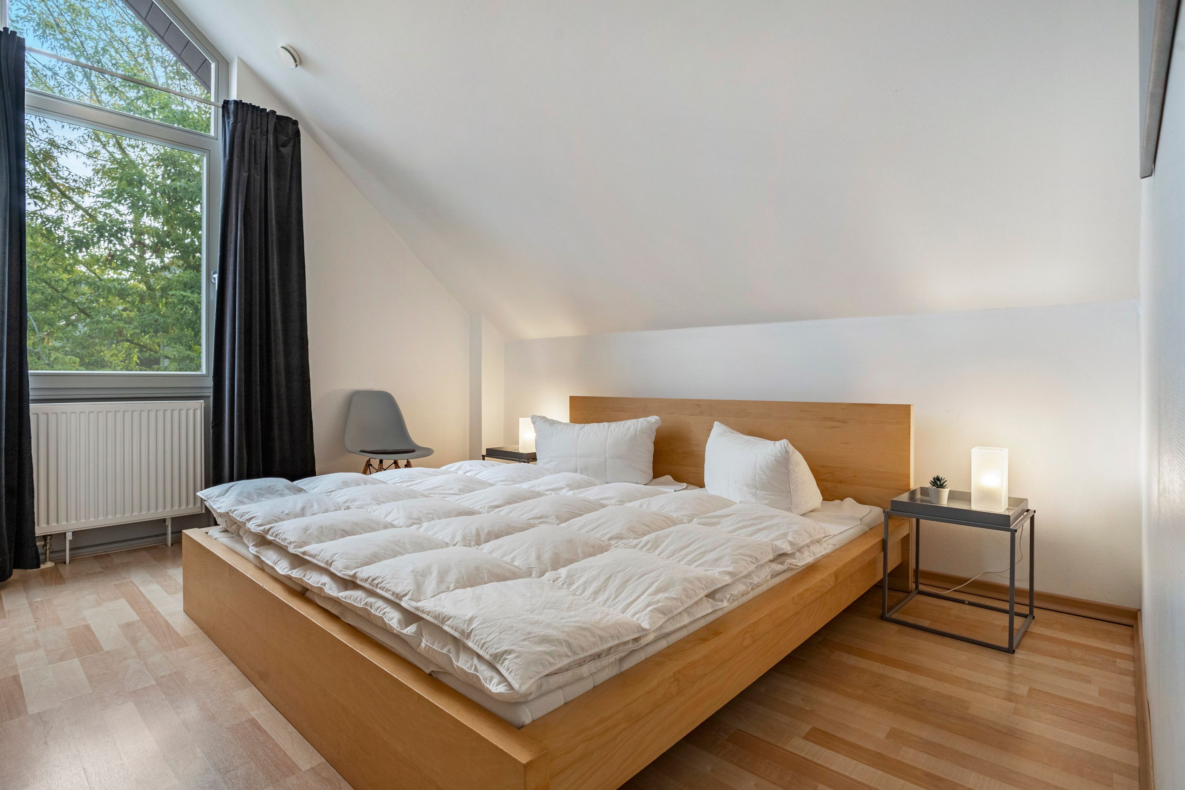 A modern bedroom with a large bed, a bedside table, a lamp, and a view of trees through a window.