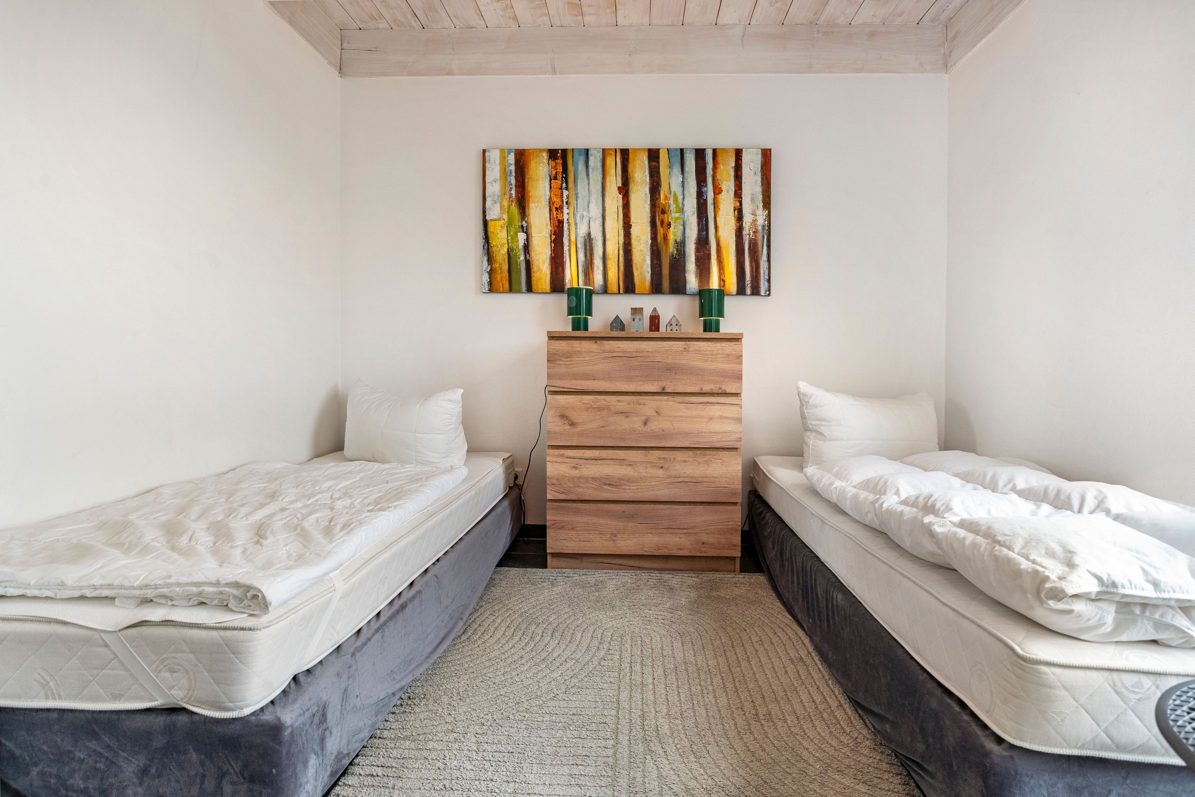 Un dormitorio con dos camas individuales, un aparador de madera, un cuadro abstracto colorido y una pequeña colección de figuritas.