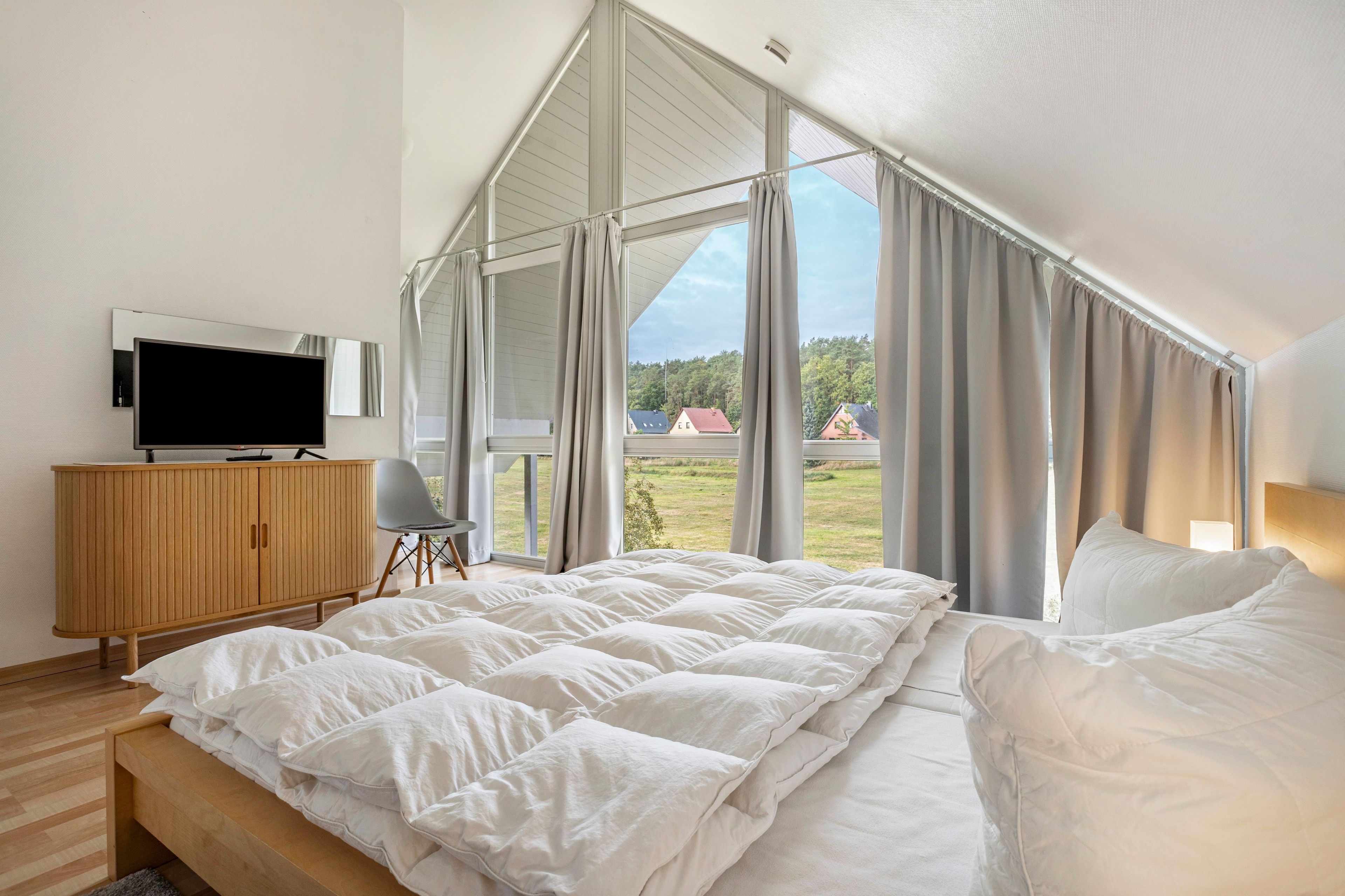 A bedroom with a large bed, a TV on a wooden cabinet, and a view of a green landscape through large windows.