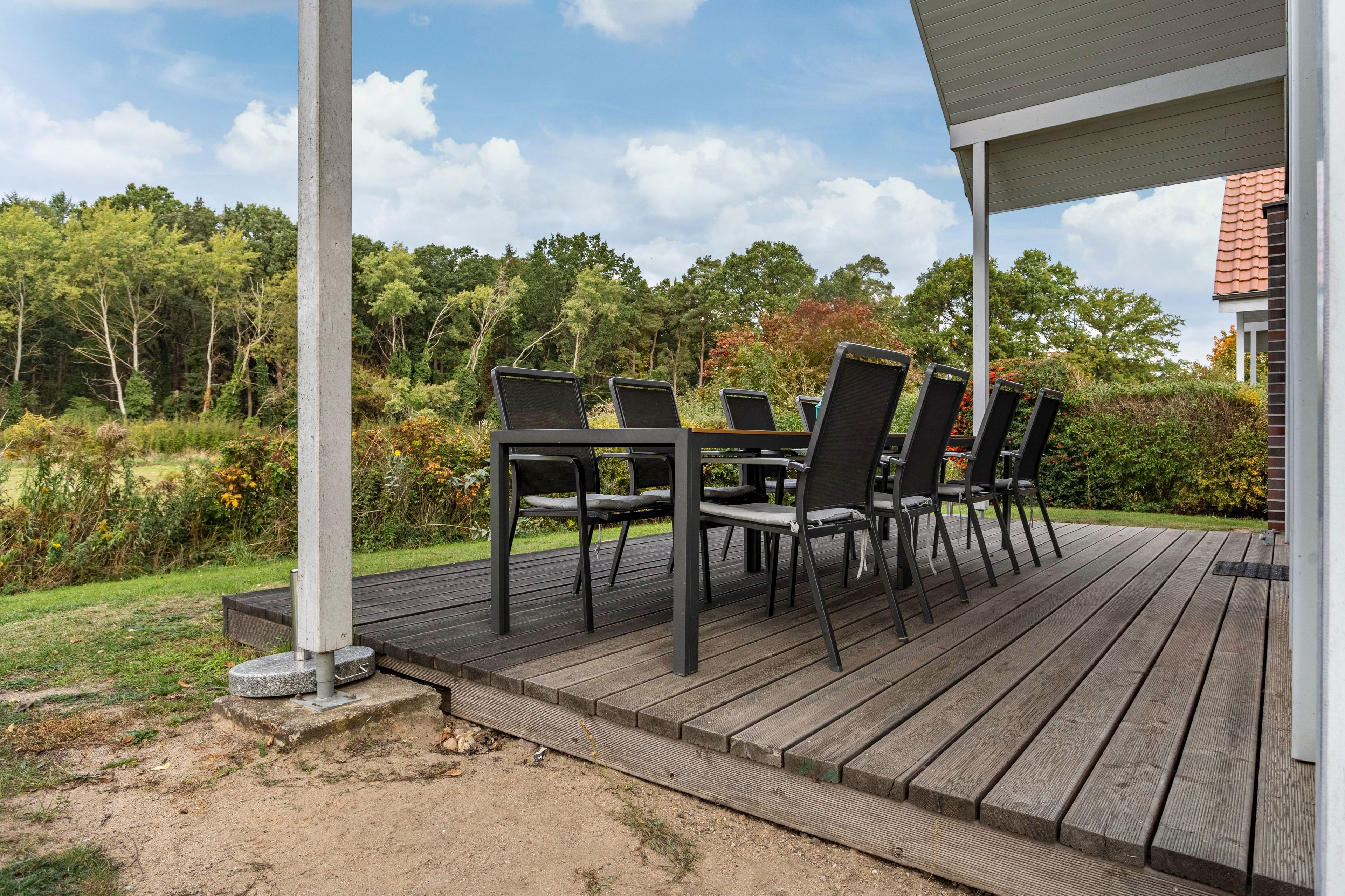 Una terraza de madera con mobiliario exterior, rodeada de follaje y un cielo despejado.