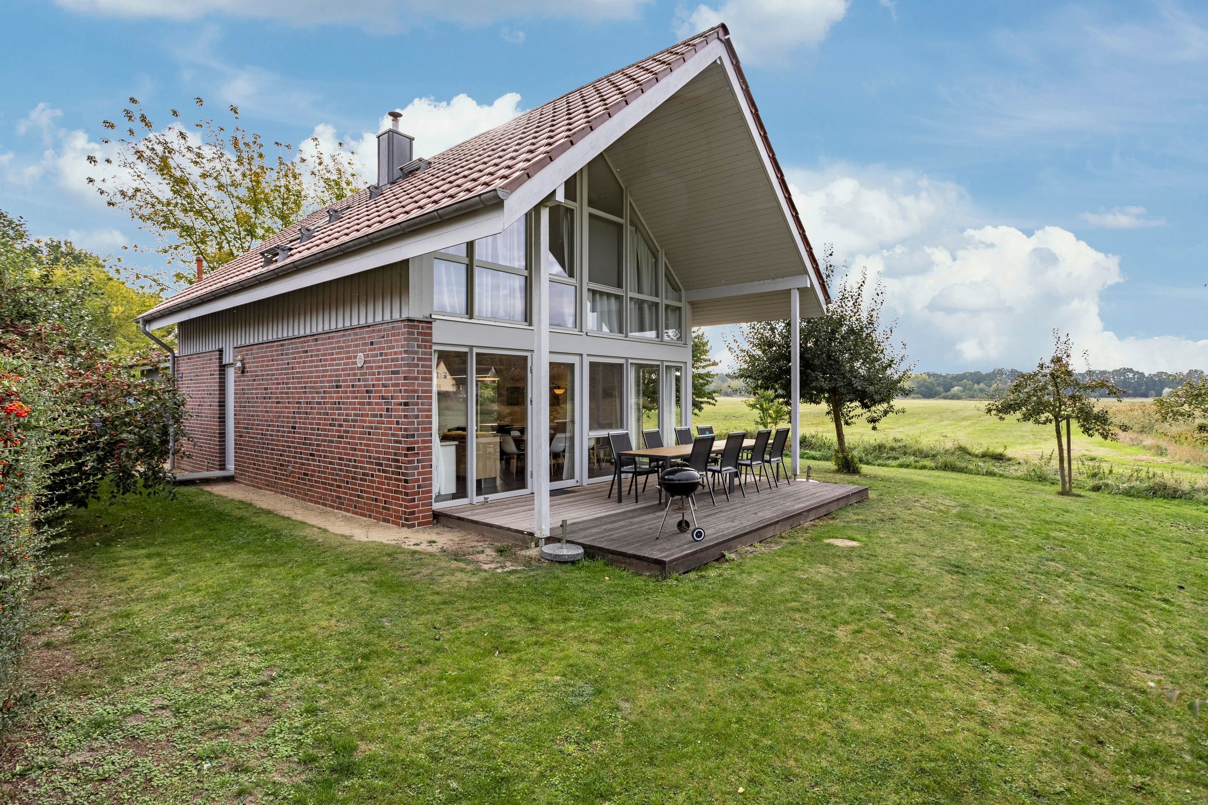 Una casa moderna de dos pisos con un amplio ventanal frontal, una terraza con mobiliario exterior y fachada de ladrillo.