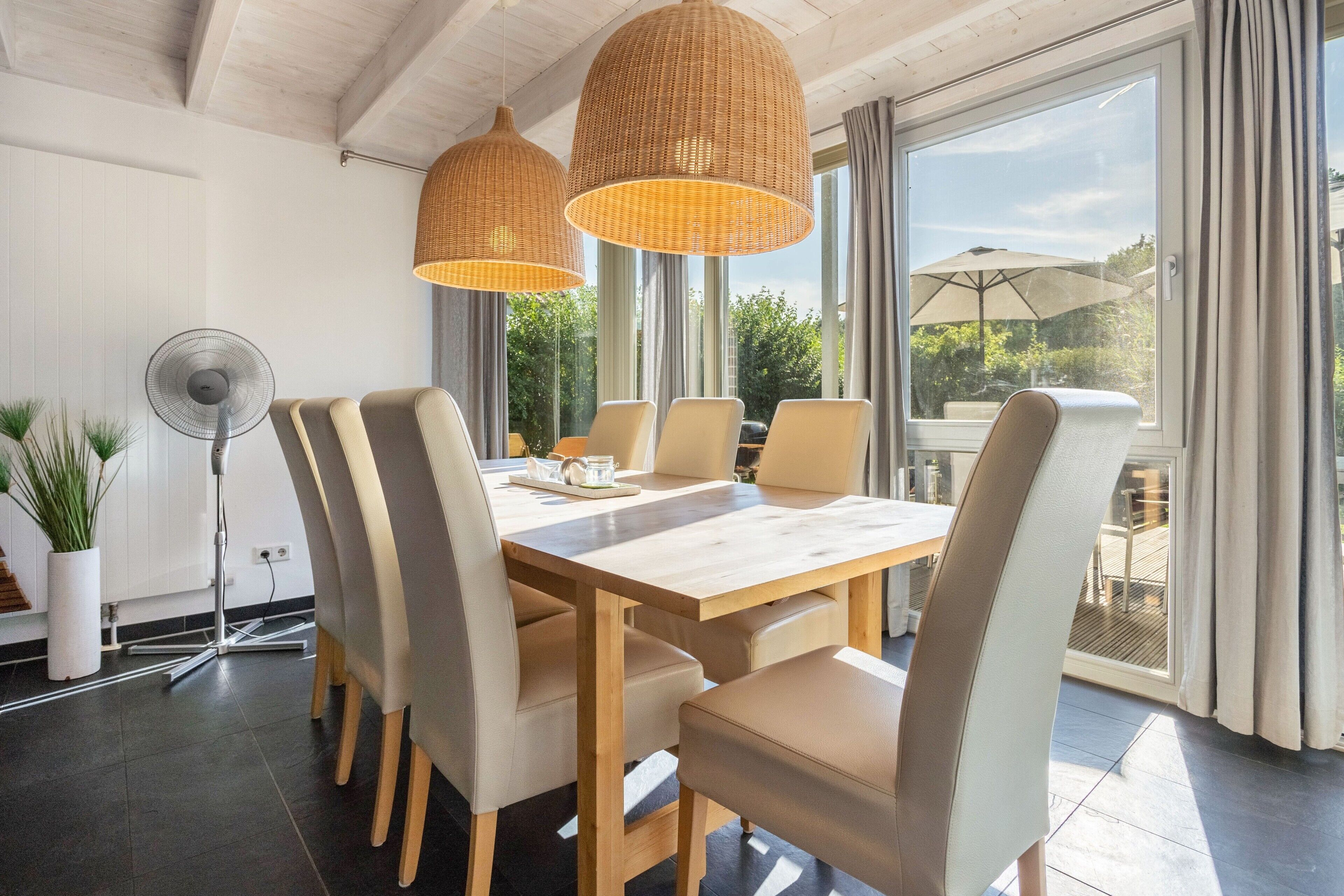A dining area with a wooden table and chairs, large windows, and a fan.
