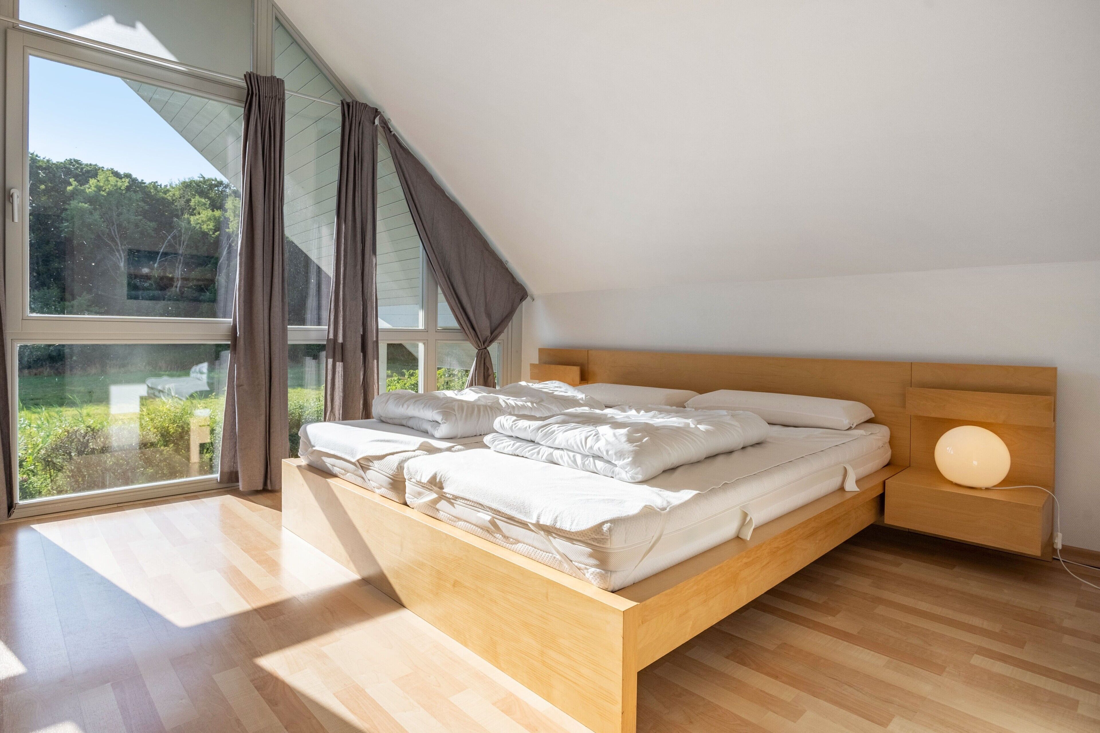 A bedroom with a wooden bed, a bedside table with a lamp, and a large window with curtains.