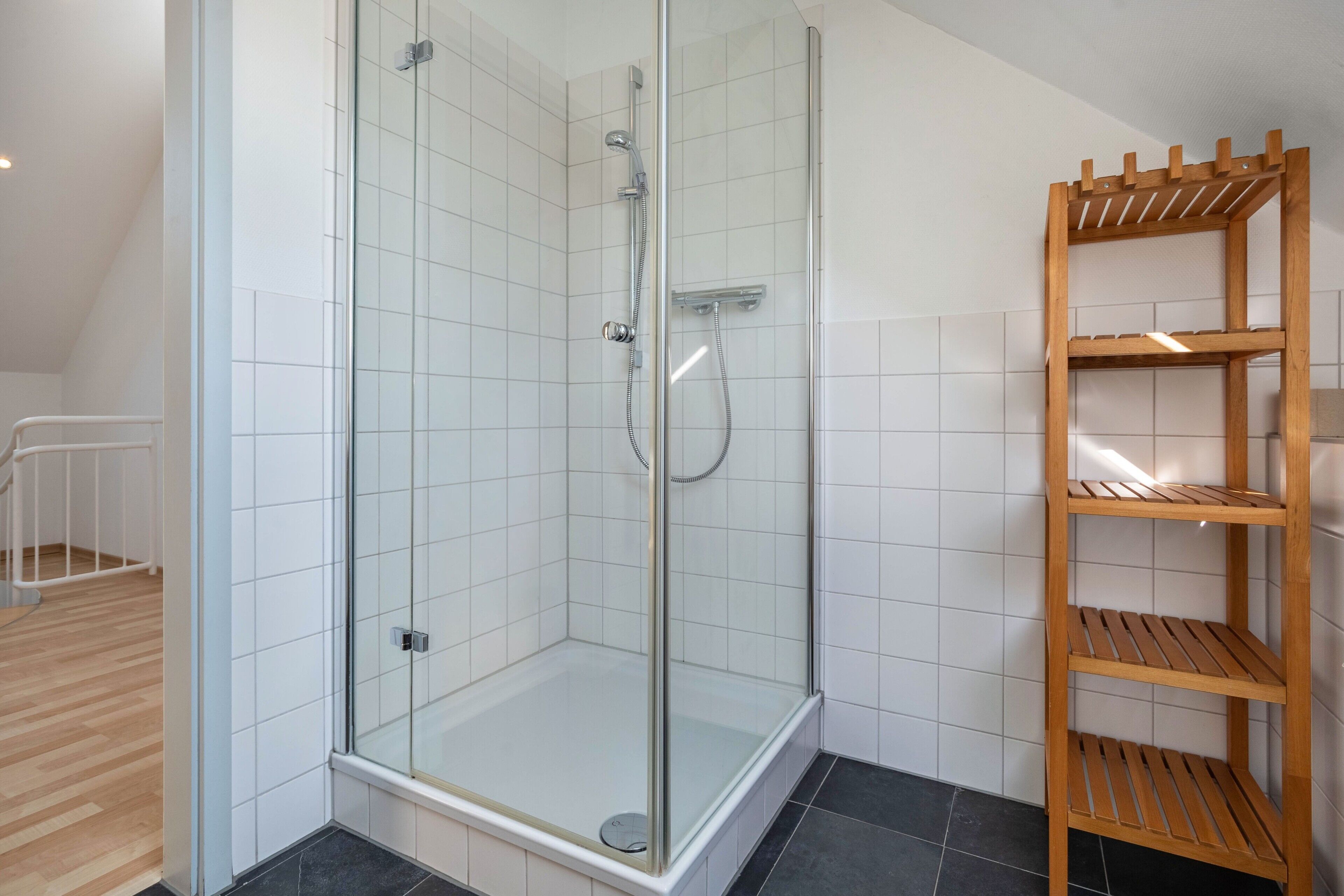 A glass-enclosed shower with a handheld showerhead, a wooden shelf unit, and a white tiled floor.