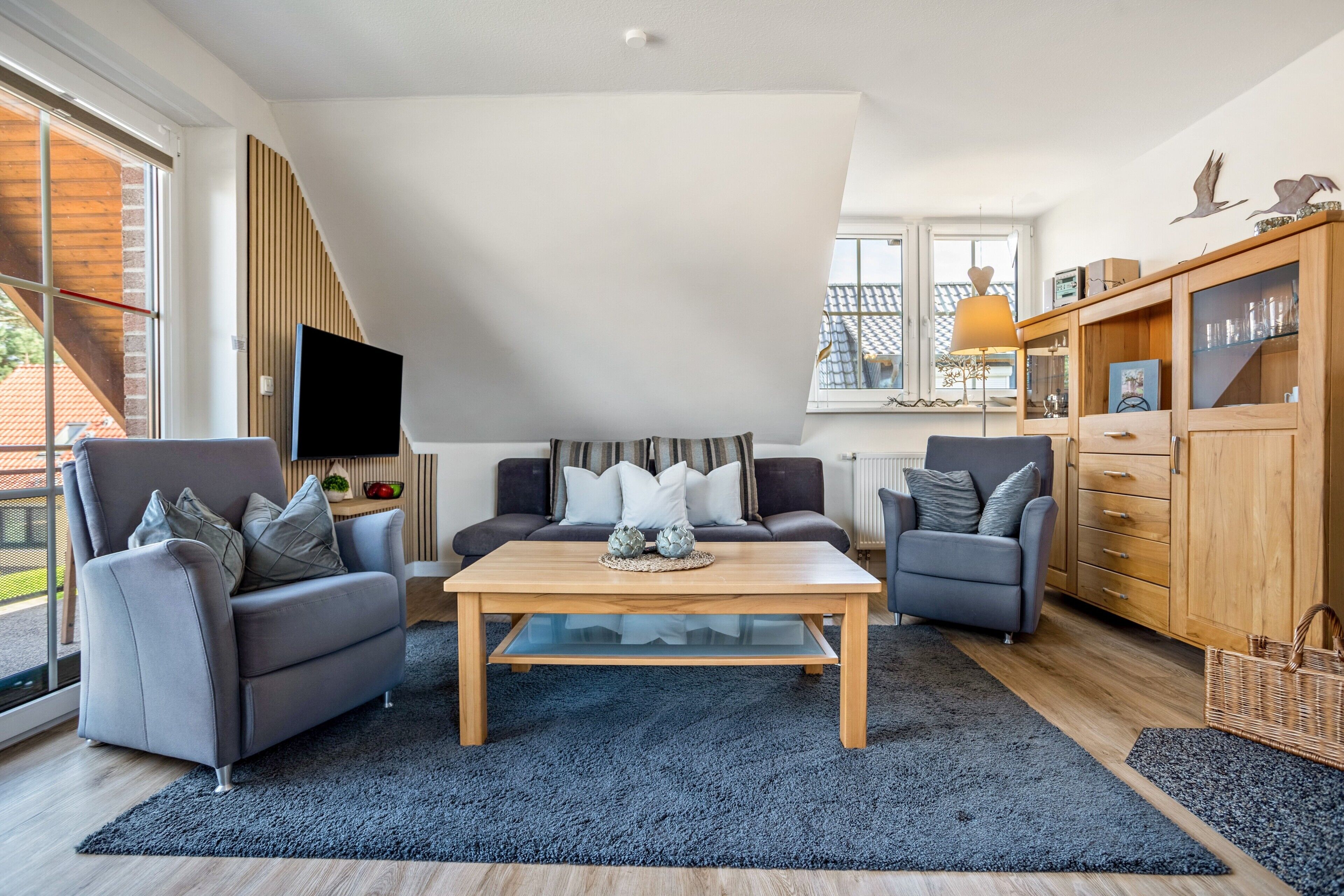 A modern living room with a high ceiling, a flat-screen TV mounted on the wall, a wooden coffee table, and two grey sofas with cushions.