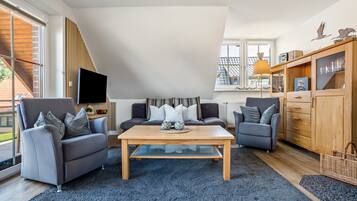 A modern living room with a high ceiling, a flat-screen TV mounted on the wall, a wooden coffee table, and two grey sofas with cushions.