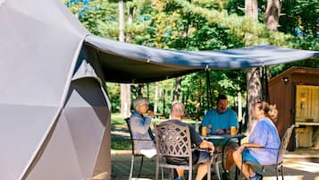 A group of people seated at an outdoor table under a tent, with a wooden structure in the background.