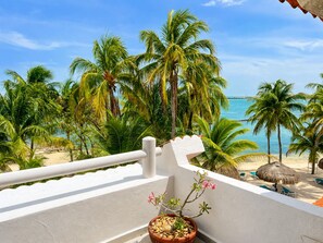 A balcony with a view of a beach, palm trees, and a thatched umbrella. - Beachfront condo with loft and sea views (Puerto Aventuras)