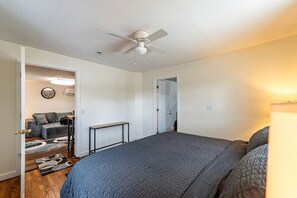 Une chambre à coucher avec un lit, un ventilateur de plafond, une porte donnant sur une autre pièce et une petite table.