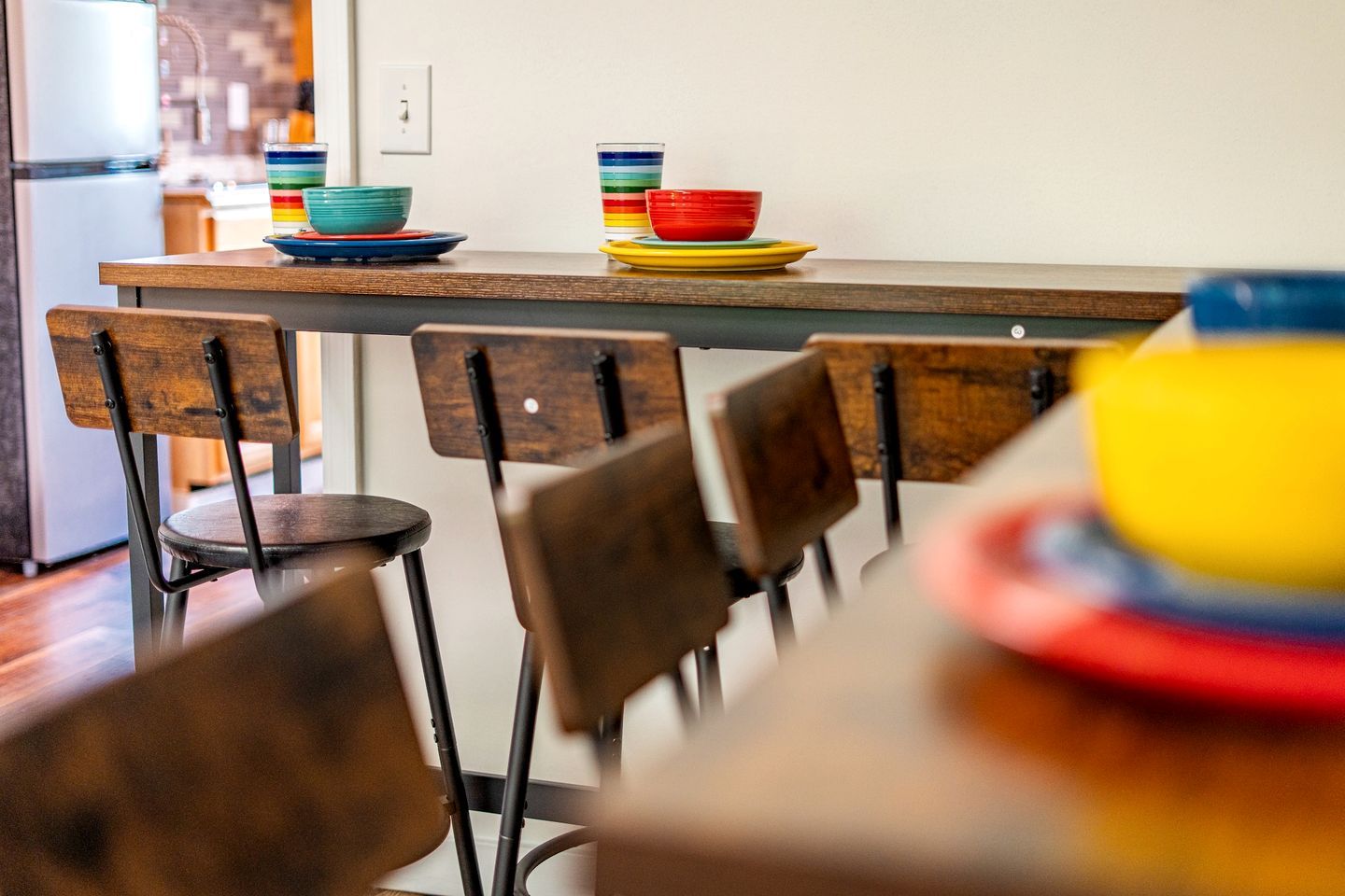 Une cuisine avec des chaises en bois, un plan de travail orné d’assiettes colorées et un réfrigérateur à l’arrière-plan.