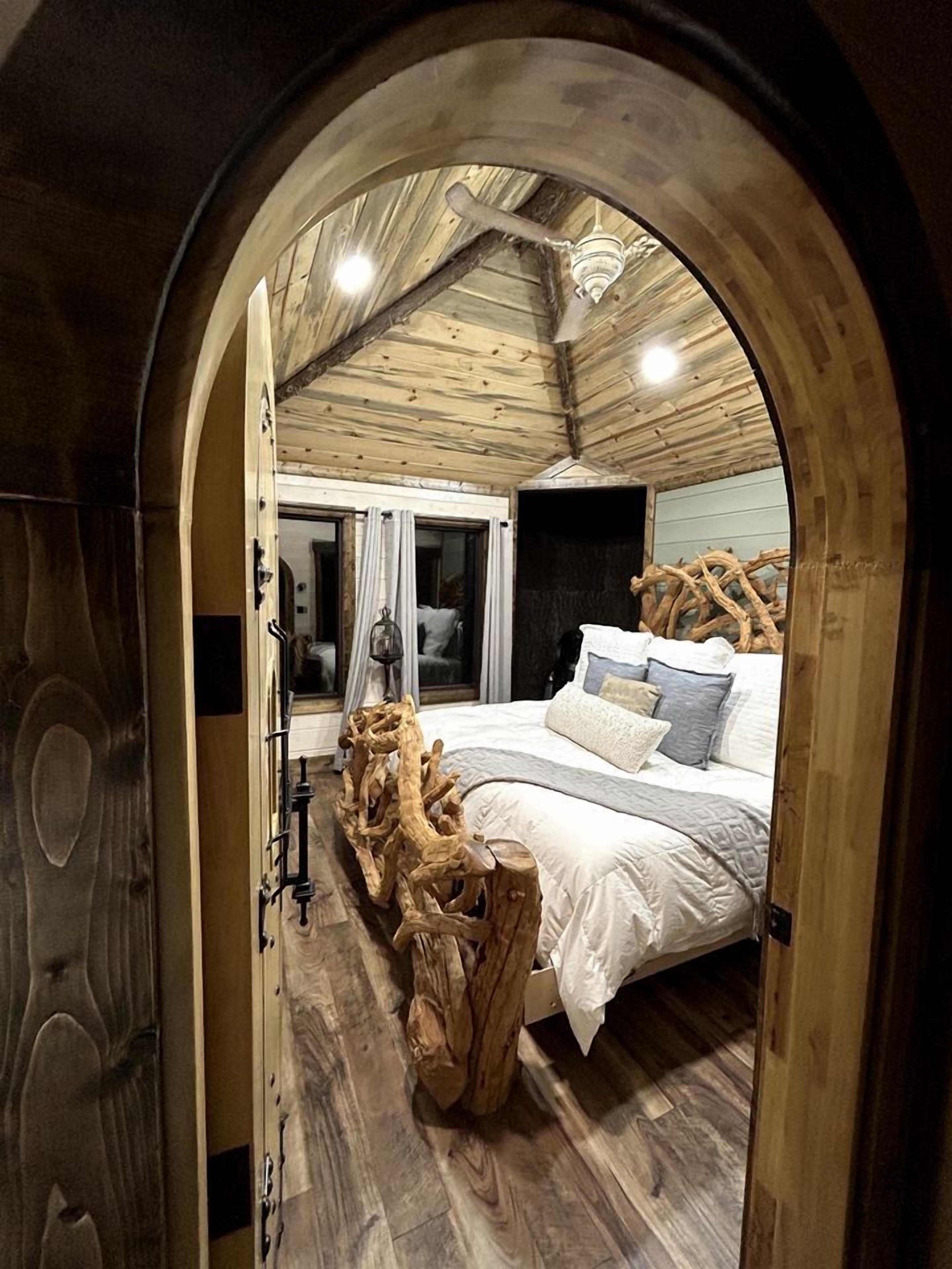A bedroom with a wooden ceiling, a bed with white bedding, and a large tree stump in the center.