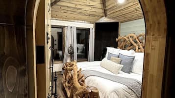 A bedroom with a wooden ceiling, a bed with white bedding, and a large tree stump in the center.