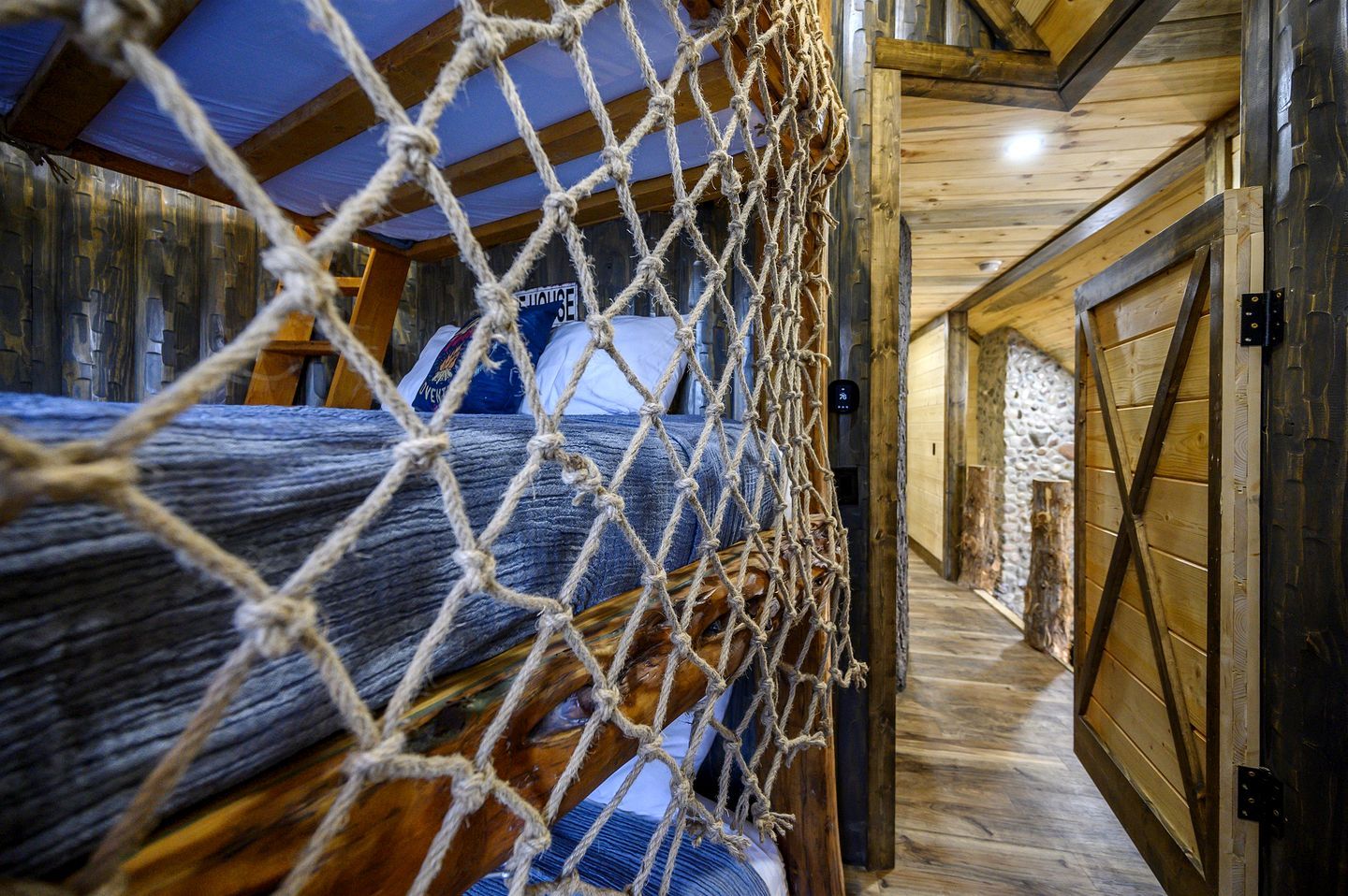 Un intérieur rustique en bois, avec une barrière faite de cordes, un lit recouvert d’une couverture bleue et un mur en pierre.