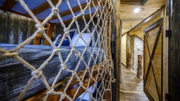 Un intérieur rustique en bois, avec une barrière faite de cordes, un lit recouvert d’une couverture bleue et un mur en pierre.