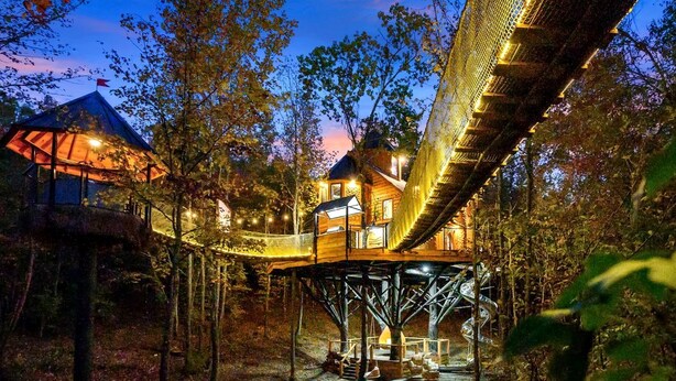 Una estructura tipo casa en un árbol con terraza de madera y zona cubierta, rodeada de árboles.