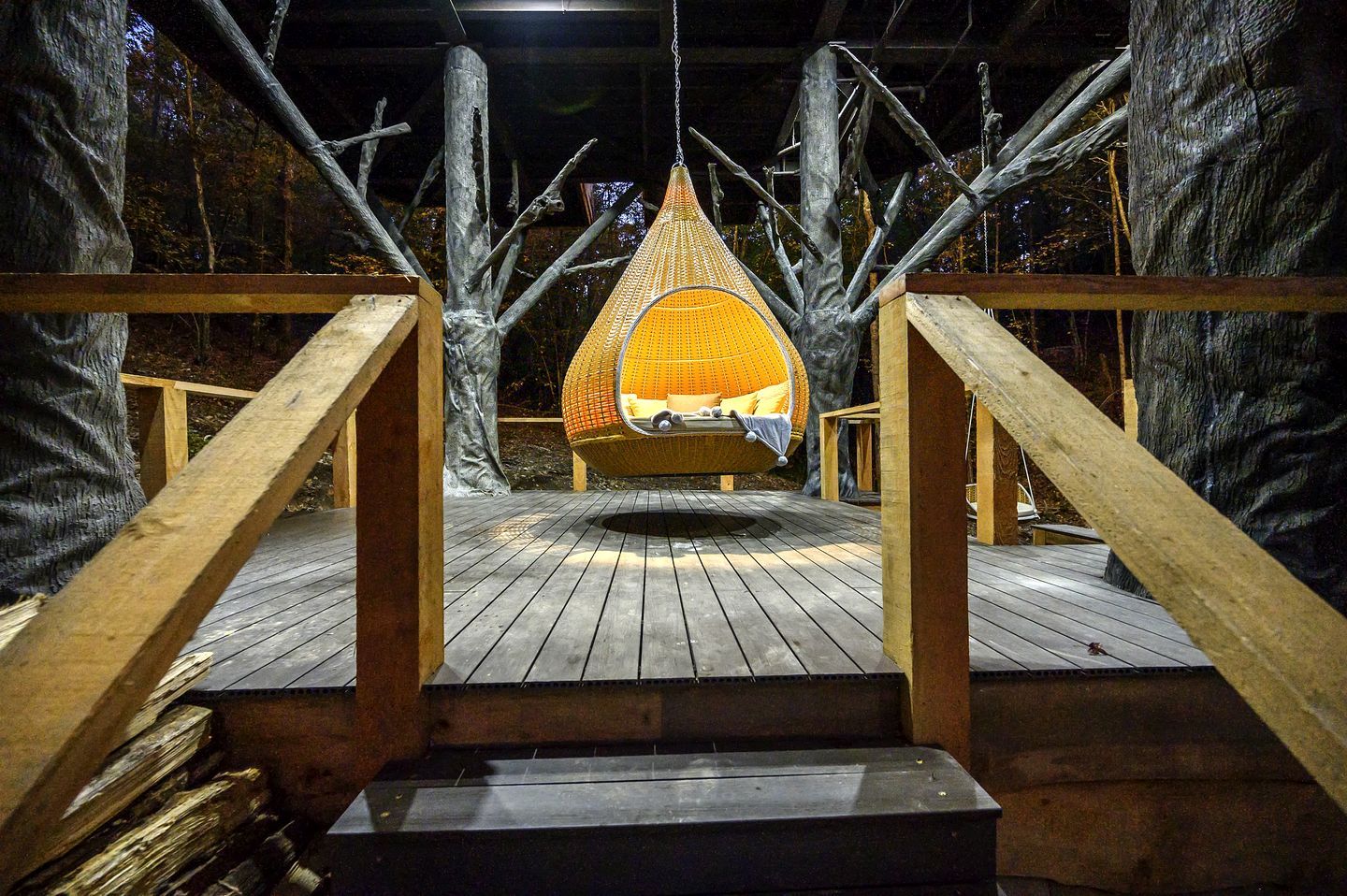 A wooden deck leading to a hanging chair with a cushion, surrounded by trees.