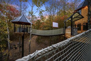 A wooden cabin with a thatched-roof structure, a hammock bridge, and a stone wall. - Gorgeous Tree House with Hot-tub in Broken Bow, Oklahoma (Broken Bow)