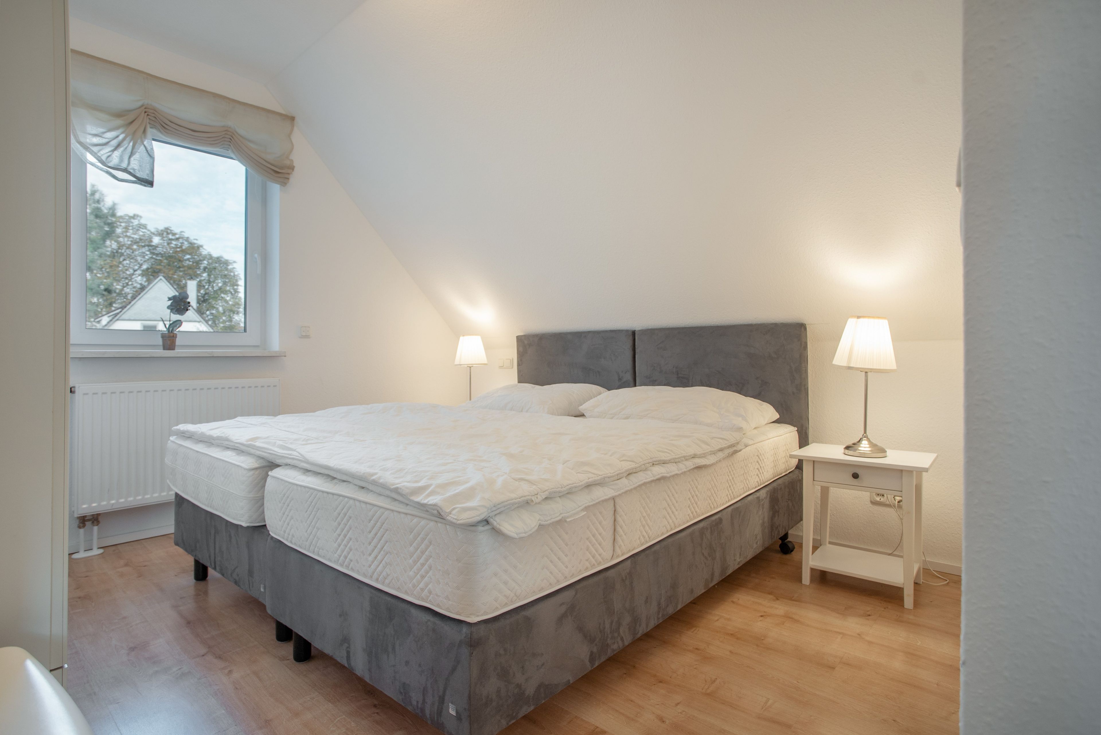 Un dormitorio con cama, lámparas de noche, una ventana con cortinas y un radiador.