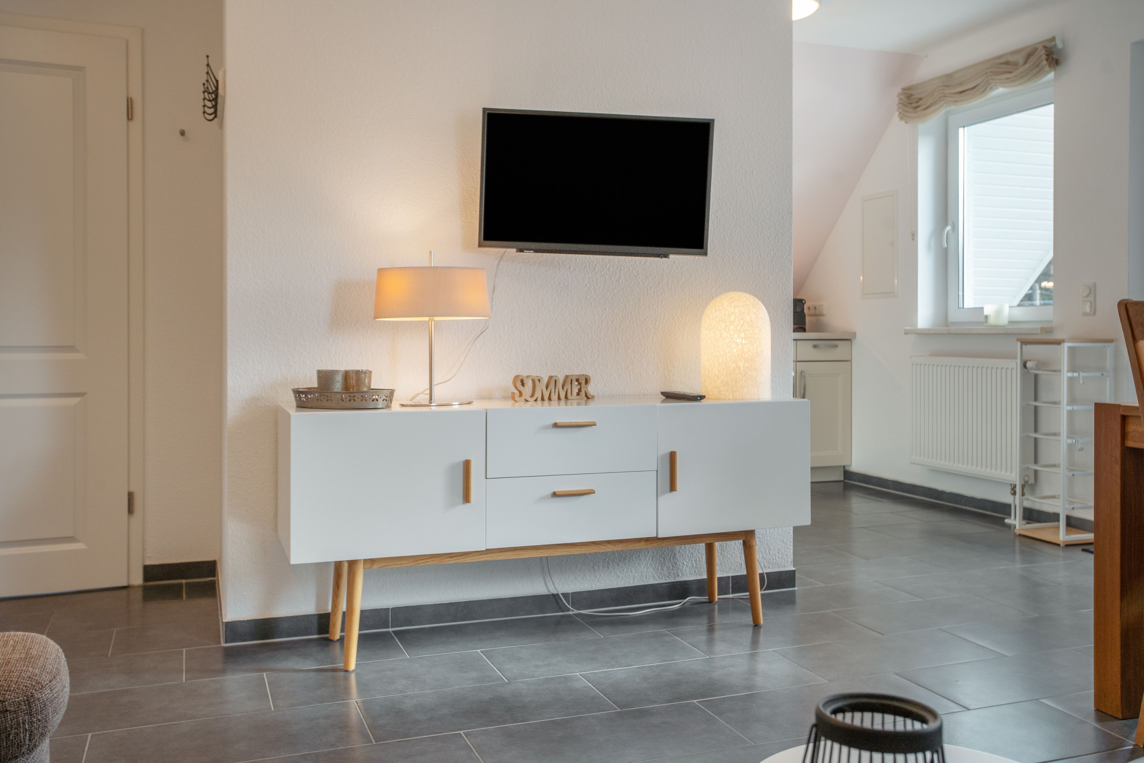 Una sala moderna con un mueble blanco, un televisor de pantalla plana montado en la pared, una lámpara y un objeto decorativo sobre el mueble.