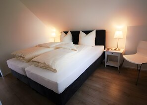 Een net opgemaakt bed met kussens, een nachtkastje met een lamp en een stoel in een moderne kamer met houten vloer.