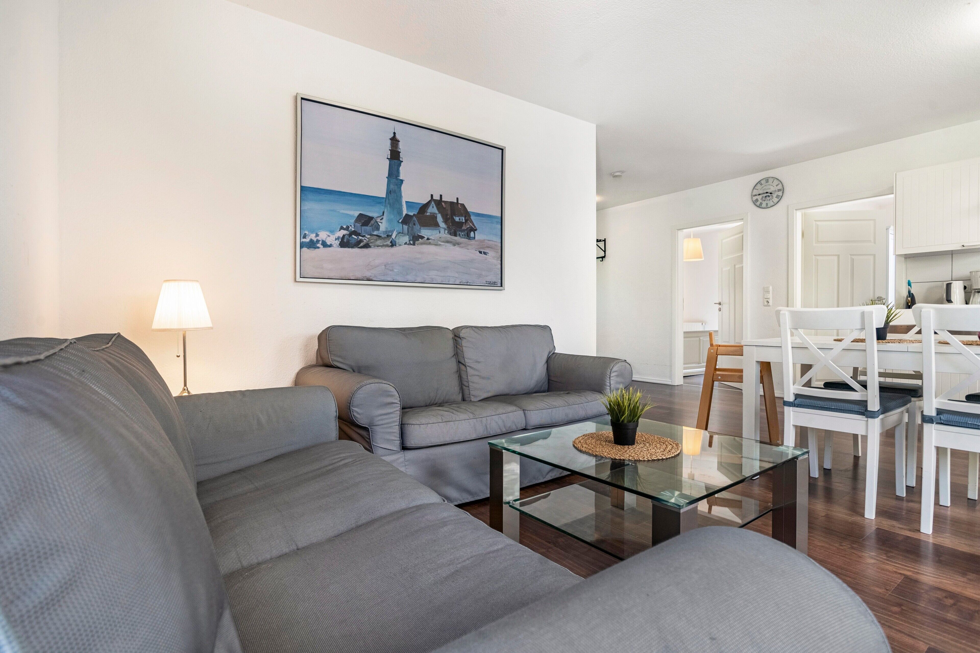 A modern living room with a grey sofa, a glass coffee table, and a painting of a lighthouse.