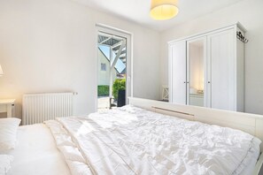 A bedroom with a large bed, a wardrobe, a radiator, and a window with a view of a house and greenery. - Apartment 'Ferienwohnung K 73 EG' with Private Terrace, Sauna, and Wi-Fi (Röbel)