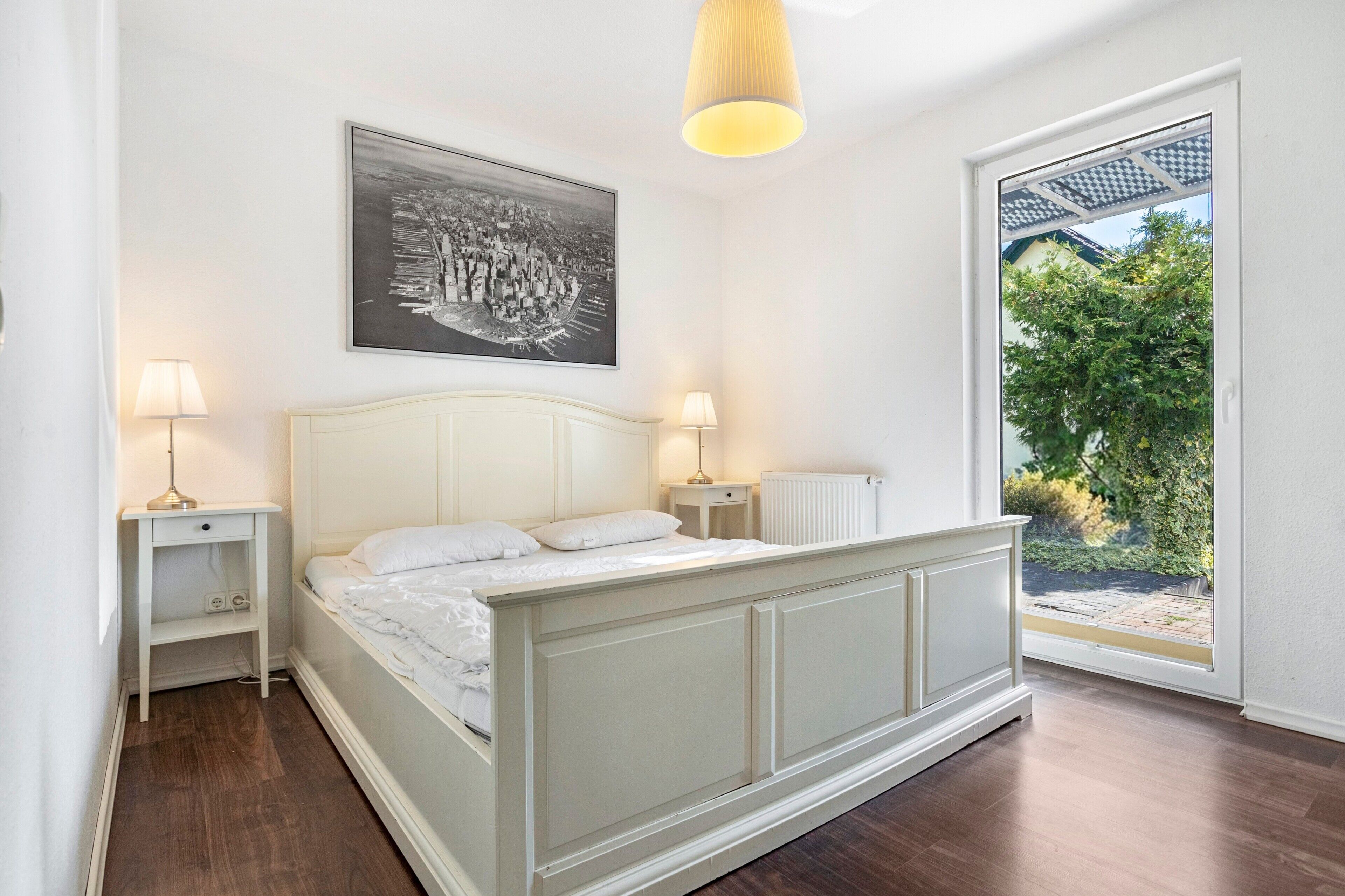 A bedroom with a bed, bedside lamps, a radiator, and a window with a view of greenery.