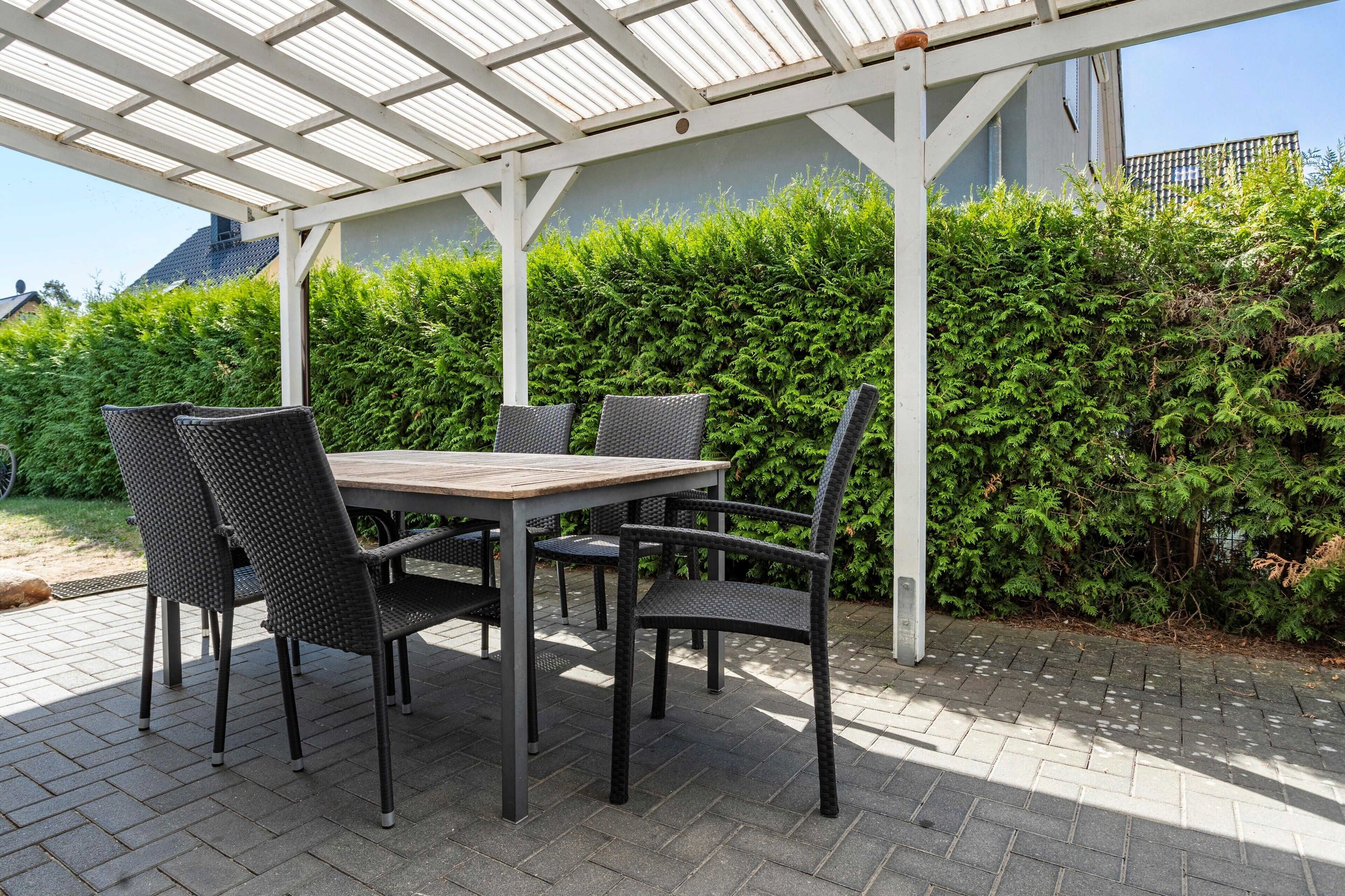 An outdoor patio with a table and chairs, a white pergola, and a hedge in the background.
