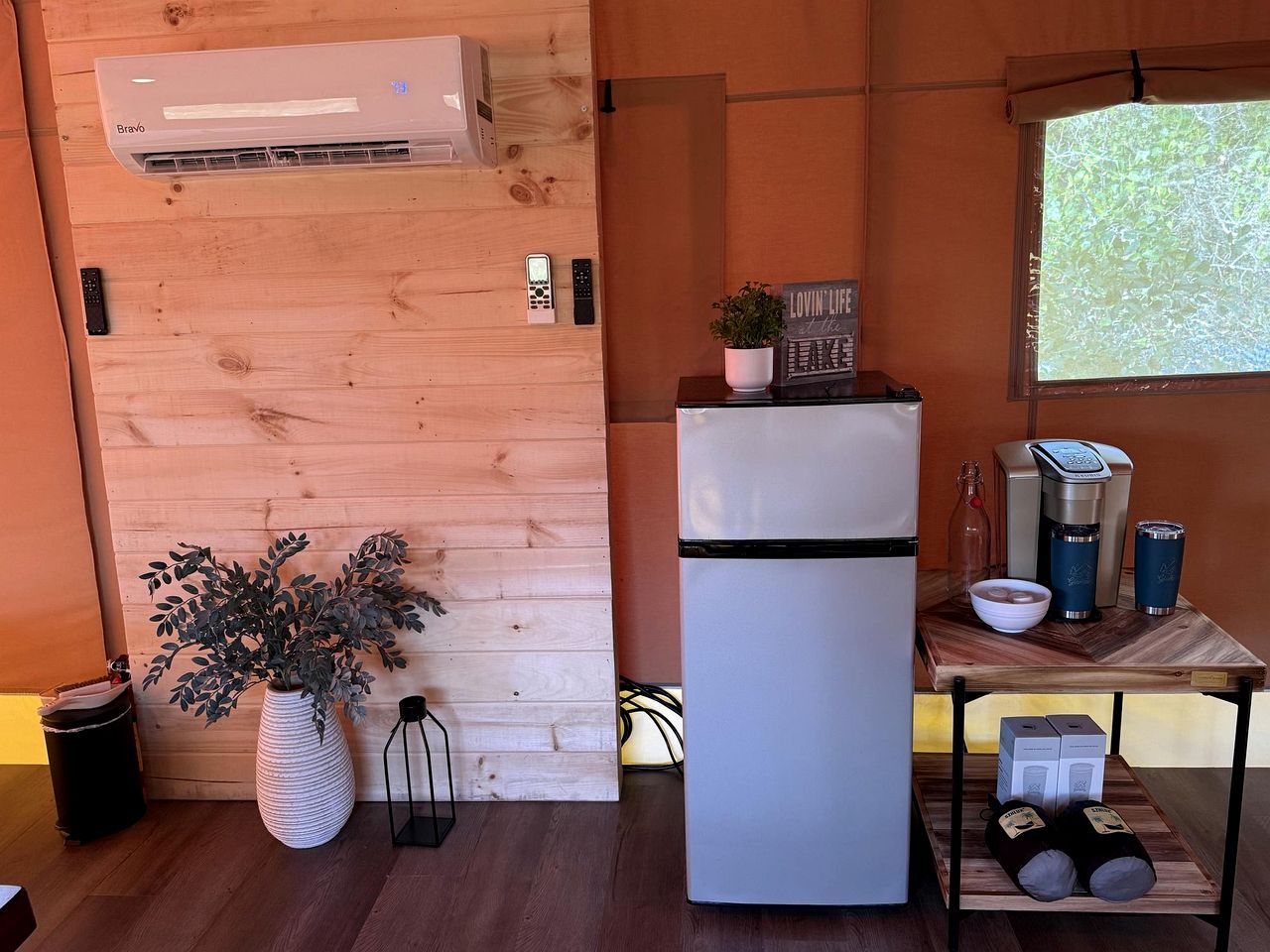 A compact kitchen with a built-in air conditioner, a refrigerator, a wooden wall, a small table with a coffee maker, and a potted plant.
