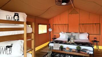 Een tent-achtige kamer met stapelbedden, een middenbed, een welkomstbord, een lamp en een kleine tafel met boeken en een plant.