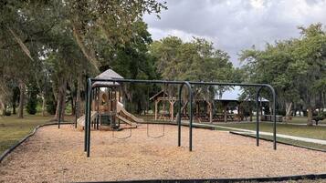 Une aire de jeux avec un toboggan, des balançoires et une structure d’escalade, le tout entouré d’arbres.